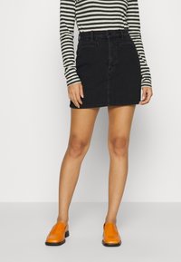 Jupe mini en denim noir à coupe ajustée, conçue avec cinq poches et une fermeture à bouton. Associée à un haut à manches longues rayé et des chaussures orange.