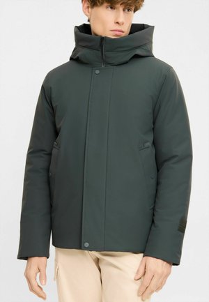 Young man wearing a dark green padded winter jacket with a high collar and front pockets, paired with light beige pants.