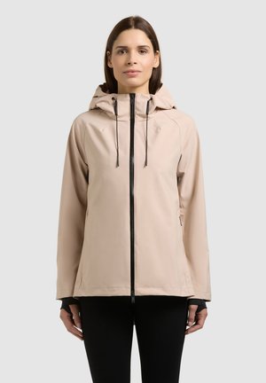 Žena oblečená v béžovej kapucňovej bunde s čiernym zipsom, stojaca pred jednoduchým svetlosivým pozadím, čelom vpred s neutrálne tvárou.