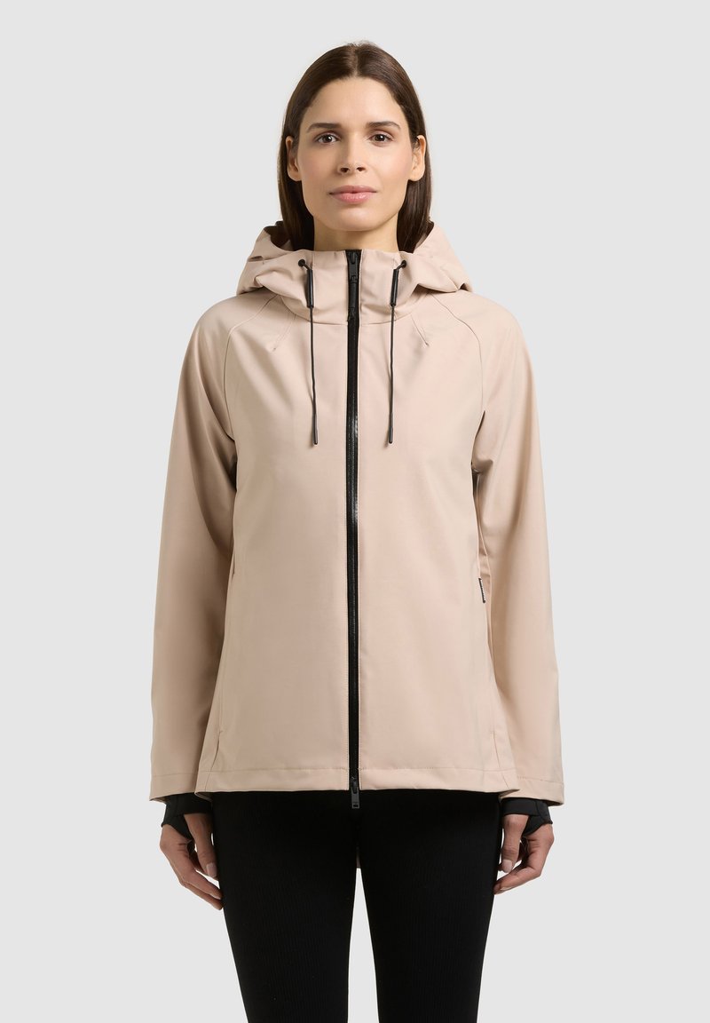 Kobieta w beżowej kurtce z kapturem i czarnym zamkiem błyskawicznym, stojąca na tle jednolitego, jasnoszarego tła, zwrócona przodem z neutralnym wyrazem twarzy.