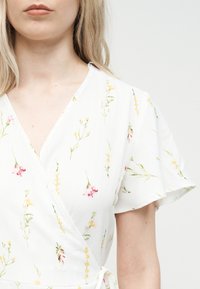 Γυναίκα που φοράει λευκό φόρεμα τύπου wrap με κοντά μανίκια και μικρό κίτρινο και ροζ ανθισμένο σχέδιο σε ανοιχτό φόντο.