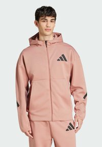 Vaaleanpunainen vetoketjullinen huppari, jossa on huppu, ja joka sisältää mustat Adidas-logot korostuksena rinnassa ja hihoissa. Huppari on valmistettu tekstuurisesta ja kevyestä kankaasta.