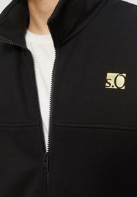 Chaqueta negra con cremallera, hecha de tela lisa, con cuello alto, dos bolsillos frontales y un pequeño logo dorado en el pecho izquierdo.