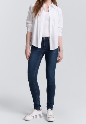 Femme portant une chemise blanche boutonnée sur un débardeur blanc, un jean skinny bleu foncé et des baskets blanches à plateforme, debout contre un fond uni.
