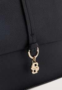 Μαύρη τσάντα από δερμάτινη υφή με λεπτό λουράκι και μικρό χρυσό μενταγιόν που απεικονίζει διαπλεκόμενα γράμματα, κρεμασμένο κοντά στο κάτω άκρο.