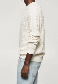 Weißer Pullover mit Kabelstrickmuster, rundem Ausschnitt und gerippten Bündchen, getragen über hellblauen Jeans, mit strukturierten Mustern im Stoff.