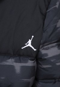 Chaqueta tipo puff negra con patrón en tonos grises, con un logo Jumpman blanco bordado en el pecho y una textura suave y acolchada.