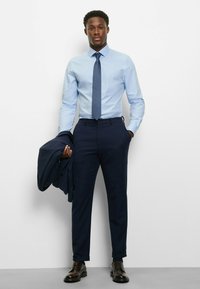 Hellblaues Hemd, gemusterte Krawatte, dunkelblaue Hose, schwarze Lederschuhe, hält einen dunkelblauen Blazer. Glatter Stoff, maßgeschneiderte Passform.