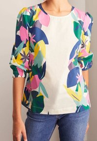 Woman wearing a white blouse with colorful abstract floral patterns on sleeves and sides, paired with blue jeans.
