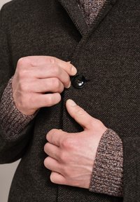 Brown herringbone wool coat with a notched collar, button closure, and textured brown sweater cuffs visible beneath.