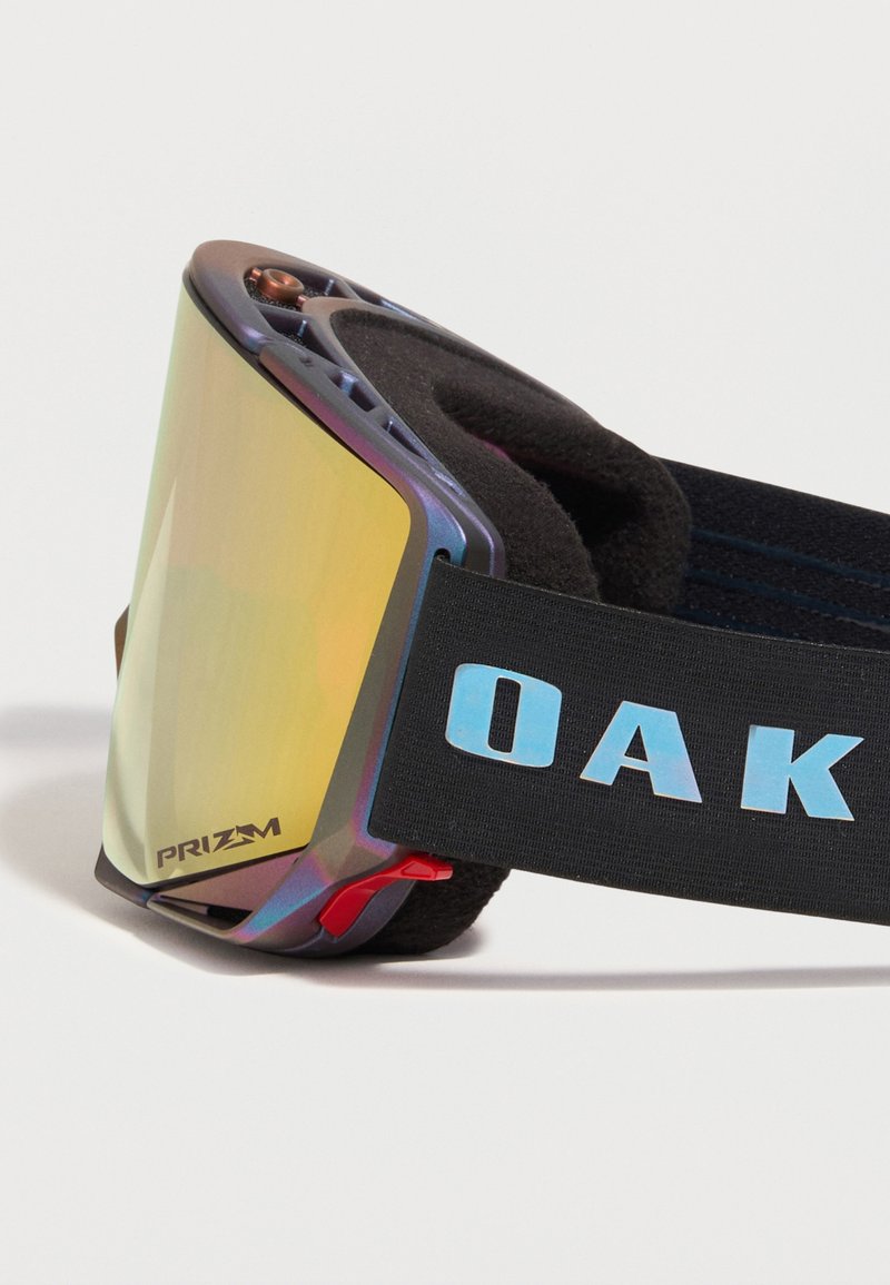 Skibril met reflecterende gele lens, zwart gevoerd montuur en zwart bandje met blauwe "OAK" tekst op witte achtergrond.