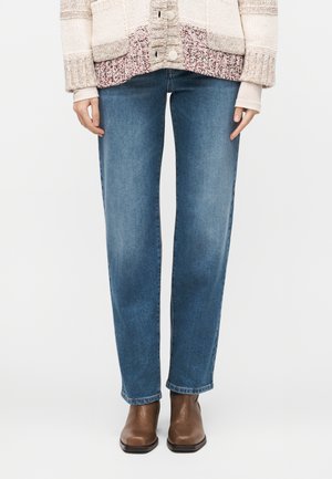 Ženska, ki nosi modre kavbojke s pravimi hlačnicami, rjave usnjene gležnarje in večbarvni pleten pulover z velikimi gumbi.