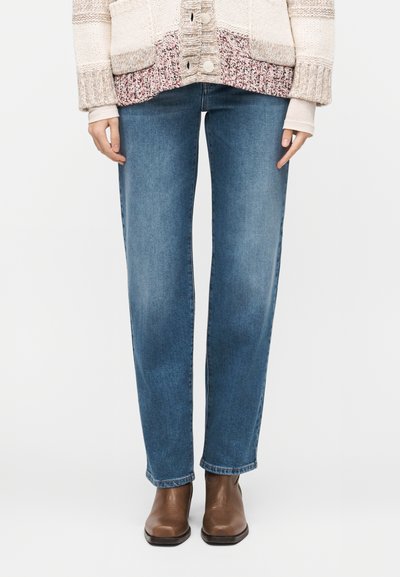 Femme portant un jean droit bleu, des bottines en cuir marron et un cardigan multicolore en tricot avec de gros boutons.