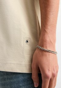 Beżowy t-shirt o teksturowanej tkaninie, z boczną metką z metalowym oczkiem. Wyeksponowany z ręką ozdobioną srebrną bransoletką.