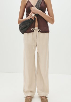 Pantalons large en crème légère avec un tissu texturé, dotés d'une taille avec cordon de serrage. Associés à un haut transparent bordeaux et un sac foncé.