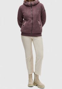 Sudadera de forro polar en un tono malva desgastado con un cuello alto, capucha con cordón, dos bolsillos frontales y puños acanalados. Combinada con pantalones cortos en blanco sucio y botas de tobillo beige.