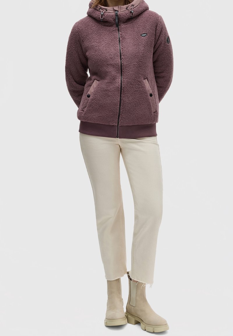 Sudadera de forro polar en un tono malva desgastado con un cuello alto, capucha con cordón, dos bolsillos frontales y puños acanalados. Combinada con pantalones cortos en blanco sucio y botas de tobillo beige.