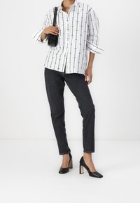 Chemise blanche boutonnée avec des lignes verticales noires et des motifs circulaires, associée à un jean slim foncé et des chaussures à talons noires.