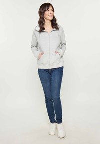 Felpa zip grigia chiara con tasche frontali, caratterizzata da una fodera interna bianca e polsini a costine, abbinata a jeans scuri e sneakers bianche.