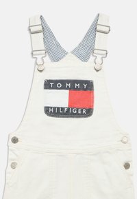 Λευκή ντένιμ φόρμα με ρυθμιζόμενους ιμάντες και μπροστινή τσέπη με αποσπώμενο λογότυπο Tommy Hilfiger σε κόκκινο, λευκό και μπλε.