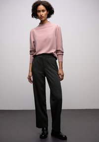 Rosa, höghalsspets med puffärmar, parat med lösa, mörka, texturerade byxor och svarta ankellånga stövlar mot en neutral bakgrund.