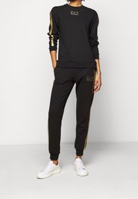 Svart sweatshirt och byxset med guldaccentstriper och logotyp. Tillverkad av mjuk tyg, med ribbade mudd och midja, kombinerat med vita sneakers.