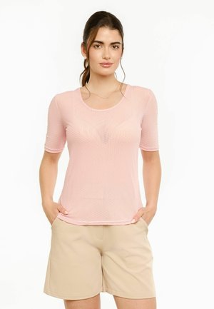 Femme aux cheveux foncés attachés en queue de cheval, portant un haut texturé rose clair à manches courtes et un short beige aux genoux, debout, les mains dans les poches.