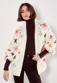 Cardigan bianco chunky lavorato a maglia con fiori ricamati colorati in rosso, arancione, rosa e verde. Presenta maniche larghe e testurizzate.