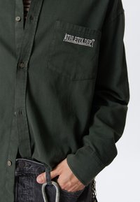 Person wearing dark green button-up shirt with "ATHLETIX.DEPT" embroidered on chest pocket, hand in black jeans pocket holding a carabiner.