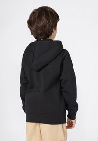 Bambino con capelli corti castani, che indossa un hoodie nero e pantaloni corti beige, rivolto di spalle contro uno sfondo chiaro e uniforme.
