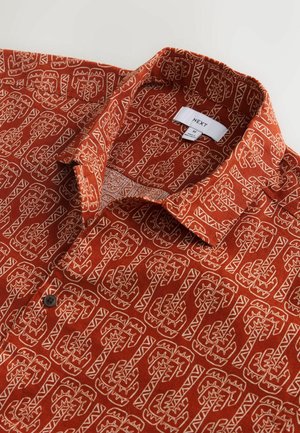 Trumpomis rankovėmis marškiniai rūdžių spalvos su balta geometrine raštu; turi apykaklę ir juodą mygtuką ant apykaklės krašto.