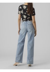 Kvinna klädd i ljusblå högmidjade vida jeans, svart blommönstrad kortärmad topp och vita klacksandaler, som står framför en grå bakgrund.