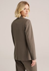Hnědý upravený blazer s elegantním designem, hladkým povrchem a dlouhými rukávy. Na zadní straně se nachází jemný šev.