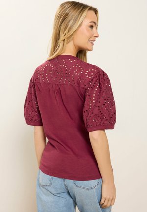 Donna vista di profilo che mostra la parte posteriore di una maglia bordeaux con maniche corte a sbuffo in pizzo traforato, abbinata a jeans celesti.