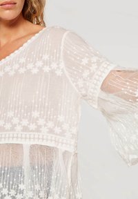 Vit broderad topp med blommönster, genomskinlig textur, utsvängda ärmar och v-ringad halsringning. Dekorativa prickiga detaljer vid fållen.