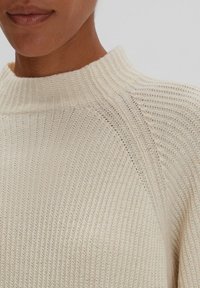 Maglione lavorato a maglia color crema con colletto alto a coste, caratterizzato da un motivo testurizzato e design a maniche raglan. Tessuto liscio e leggero.