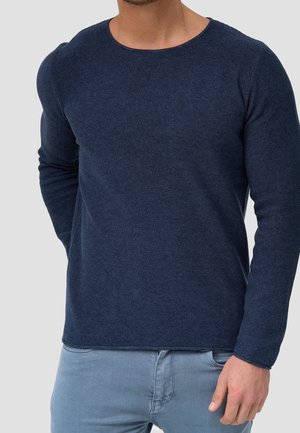 Man wearing dark blue textured long-sleeve shirt and light blue jeans, standing with one hand near pocket against plain background.