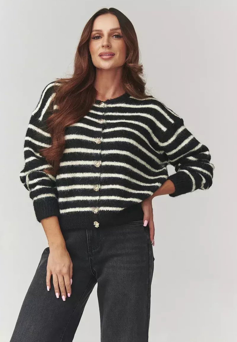 Kobieta z długimi brązowymi włosami, ubrana w sweter w czarno-białe paski z guzikami i czarne dżinsy, pozująca z jedną ręką na biodrze na jednolitym tle.