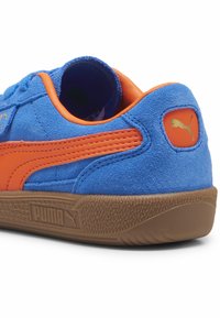 Mėlynos zomšinės sportinės krepšinio bateliai su oranžiniais akcentais, turintys tekstūruotą gumos padą ir logotipą šalia kulno. Paprastas, sportiškas dizainas.