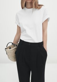 Vit t-shirt med rullade ärmar som matchas med högmidjade svarta plisserade byxor; håller en liten vävd korgväska med svarta handtag.