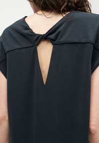 Γυναίκα που φοράει μαύρο τοπ με στριφτό κόμπο και τριγωνικό άνοιγμα στην άνω πλάτη, δείχνοντας γυμνό δέρμα και σγουρά μαλλιά.