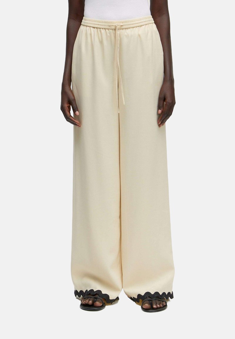 Brede crèmekleurige broek met een elastische tailleband, voorzien van een trekkoord en zwarte golvende bies aan de onderkant, gemaakt van een zachte stof.