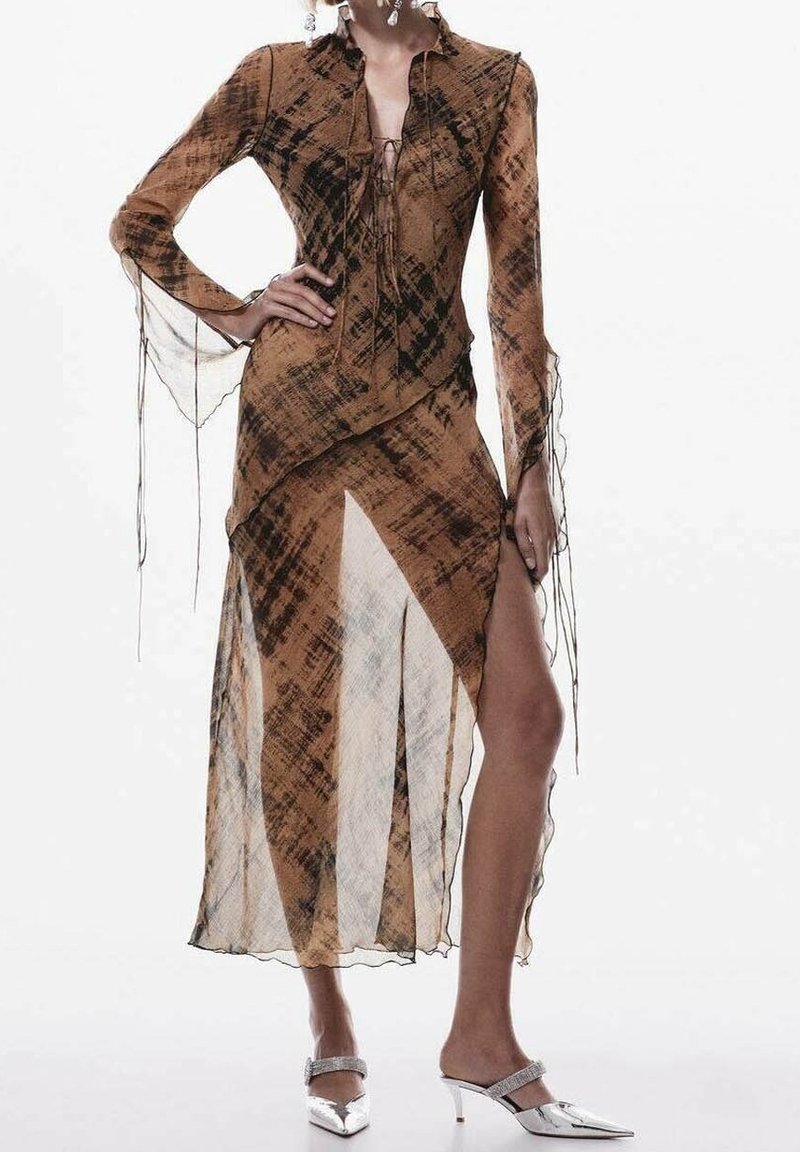 Femme portant une robe transparente marron et noire à motifs, manches longues, fente haute, et talons aiguilles argentés.