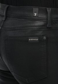 Közeli kép fekete farmernadrág hátulzsebéről, amelyen "for all mankind" feliratú hímzett címke és egy kis szövetcímke látható a 7-es számmal a derékpánton.