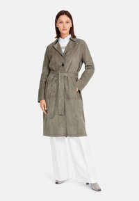 Suede trench coat in olive green, featuring a single button closure, belted waist, and side pockets. Paired with wide white pants and gray shoes.