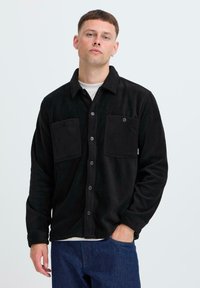 Black fleece shirt jacket with two front pockets, button closure, and a collar. Features a soft texture and relaxed fit. Worn over a white top.