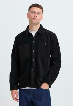 Zwarte fleece shirtjas met twee voorzakken, knoopsluiting en een kraag. Kenmerkt zich door een zachte textuur en een ontspannen pasvorm. Draagt over een witte top.