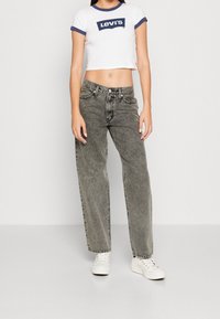 Joven mujer vestida con una camiseta corta blanca de Levi’s, jeans gris de lavado ácido de talle alto y zapatillas blancas, de pie frente a un fondo blanco.