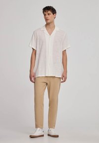 Camicia bianca di lino a maniche corte con bottoni frontali, abbinata a pantaloni tapered beige chiaro e sneaker bianche.