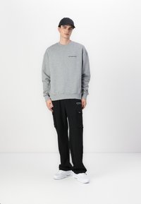 Sudadera gris con texto, pantalones cargo negros con bolsillos laterales y zapatillas blancas. El modelo lleva una gorra negra, frente a un fondo liso.